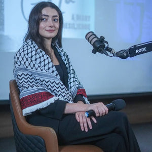Woman sitting in front of a microphone with a Rode brand name, wearing a patterned shawl.